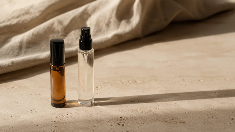 A small amber dab-on oil bottle beside a slim clear-glass eau de parfum spray on travertine, long shadows — two ways a scent can wear.