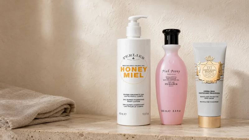 Honey Miel body lotion, Pink Peony shower gel and Imperial Honey hand cream grouped on a travertine shelf in morning light — a small scent wardrobe.
